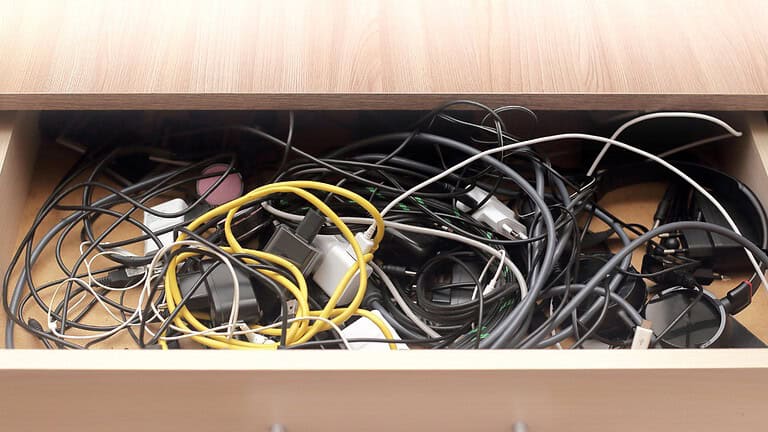 Messy drawer filled with tangled cables, chargers, and power adapters, various electronic cords mixed together, unorganized storage of wires, cluttered tech accessories in a wooden drawer
