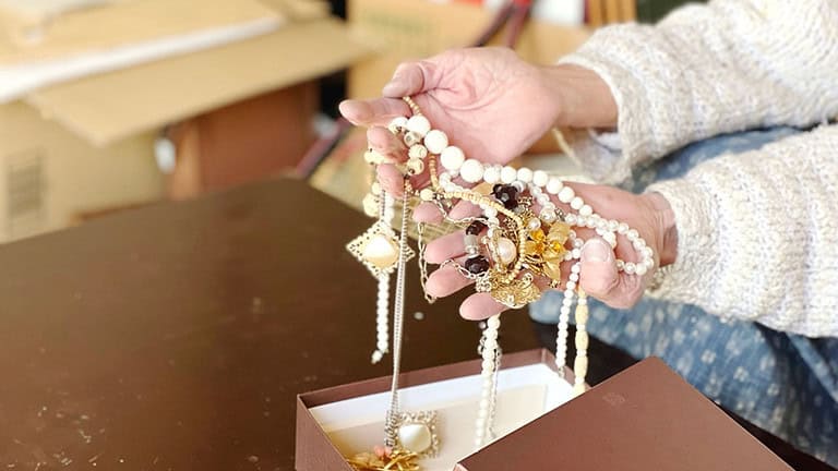 A hand holding multiple pieces of jewelry, including necklaces and beaded accessories, displayed over a small stand or platform, likely in a craft or retail setting