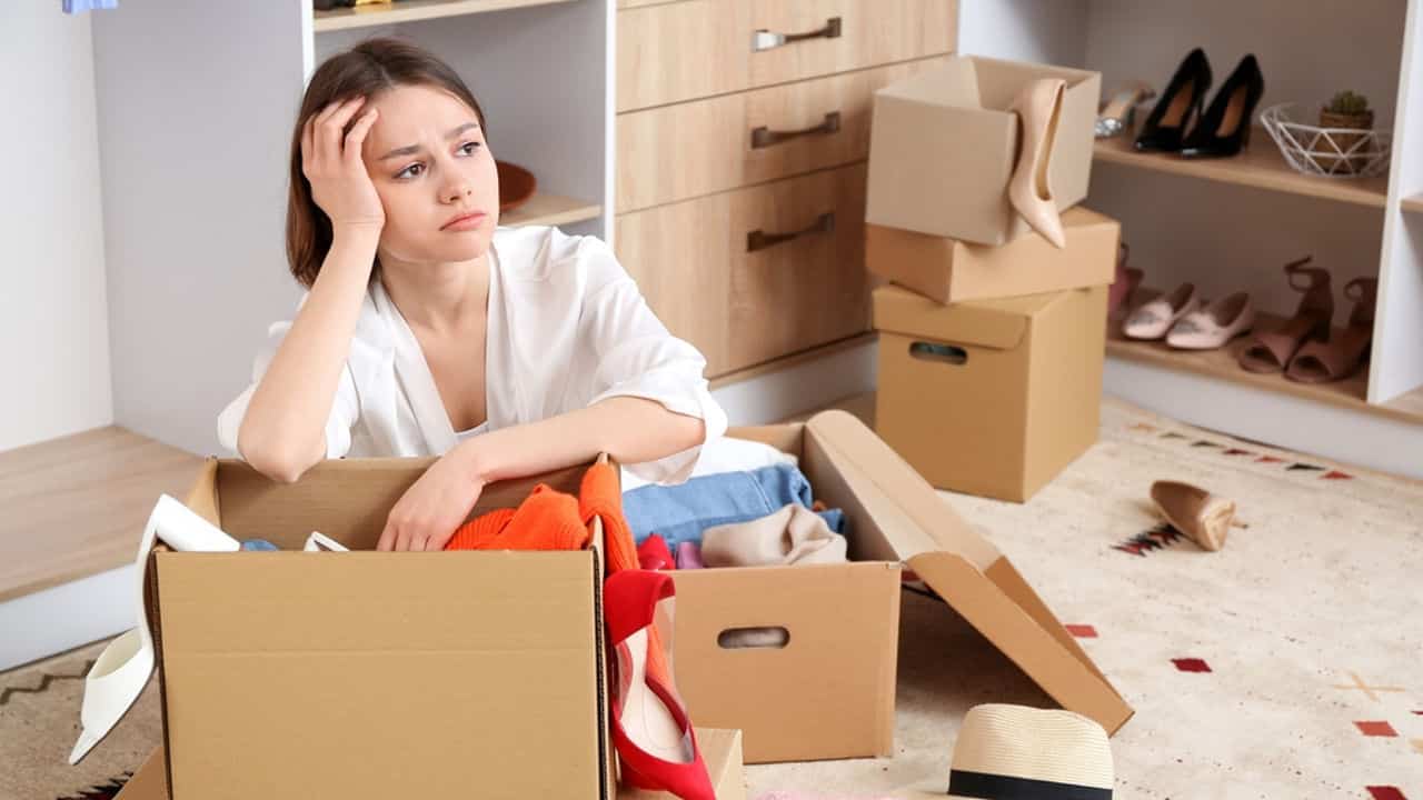 Young woman sitting on the floor surrounded by open cardboard boxes, resting her head on her hand with a tired expression, packing or unpacking clothes and shoes in a bedroom, indicating stress or exhaustion during a move or room reorganization