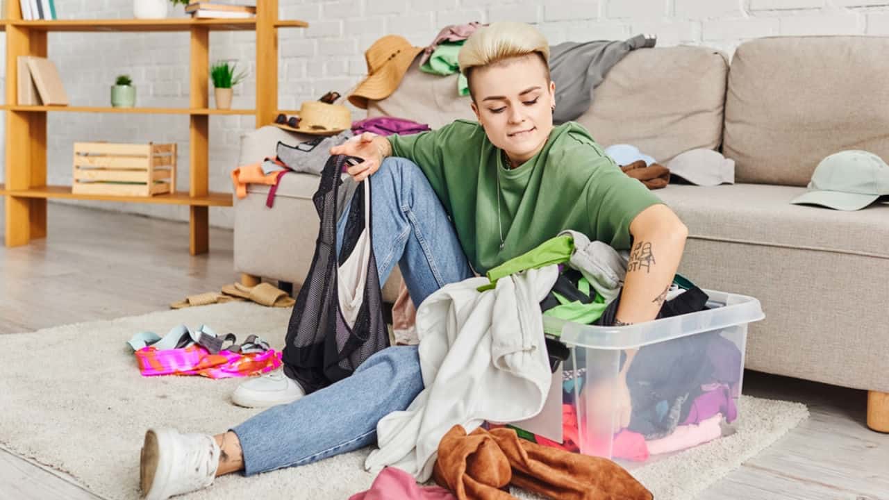 Person sorting through a pile of clothes, cluttered room with scattered items, organizing wardrobe, decluttering space, folding and placing clothes in a storage bin, shoes and hats in the background, home cleaning effort
