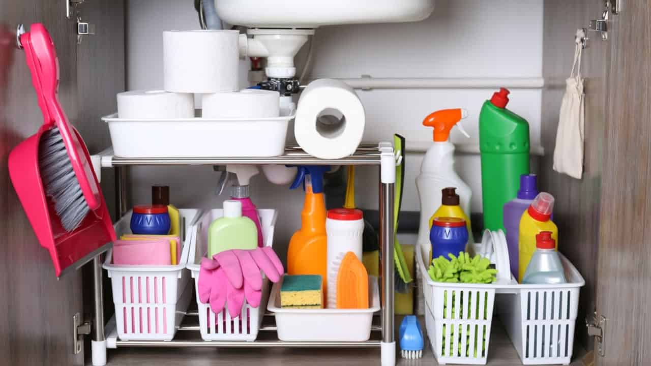 Neatly arranged cleaning supplies, plastic baskets holding sponges, brushes, and gloves, various bottles of detergents and sprays, paper towels and toilet rolls stored conveniently, efficient home cleaning organization