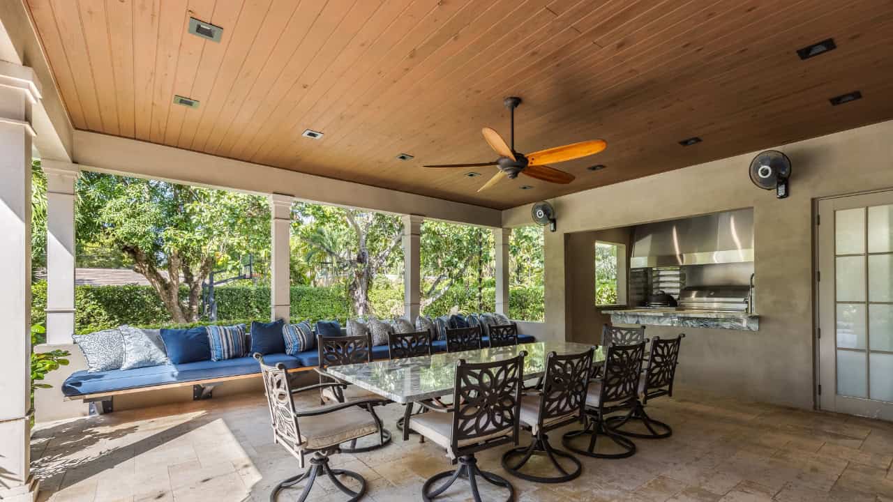Covered outdoor patio with dining table, chairs, ceiling fan, wood ceiling, large windows overlooking greenery, tile flooring