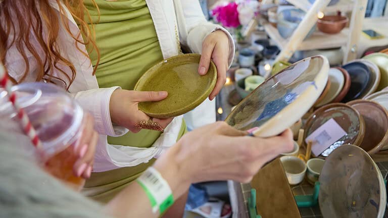 Hands examining a green striped fabric item while thrift shopping, with other secondhand clothing and textiles visible in the background at a thrift store or vintage shop