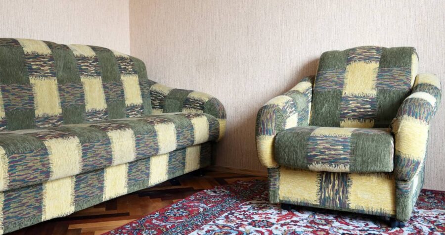 Living room corner with two patterned sofas or armchairs, decorative area rug with red and blue pattern, white wall, blue checkmark icon in upper right corner.