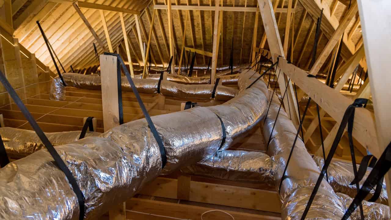 Ductwork in an attic, insulated pipes suspended from the ceiling, wooden beams supporting the structure, air ducts secured with straps, attic space with visible roof slats