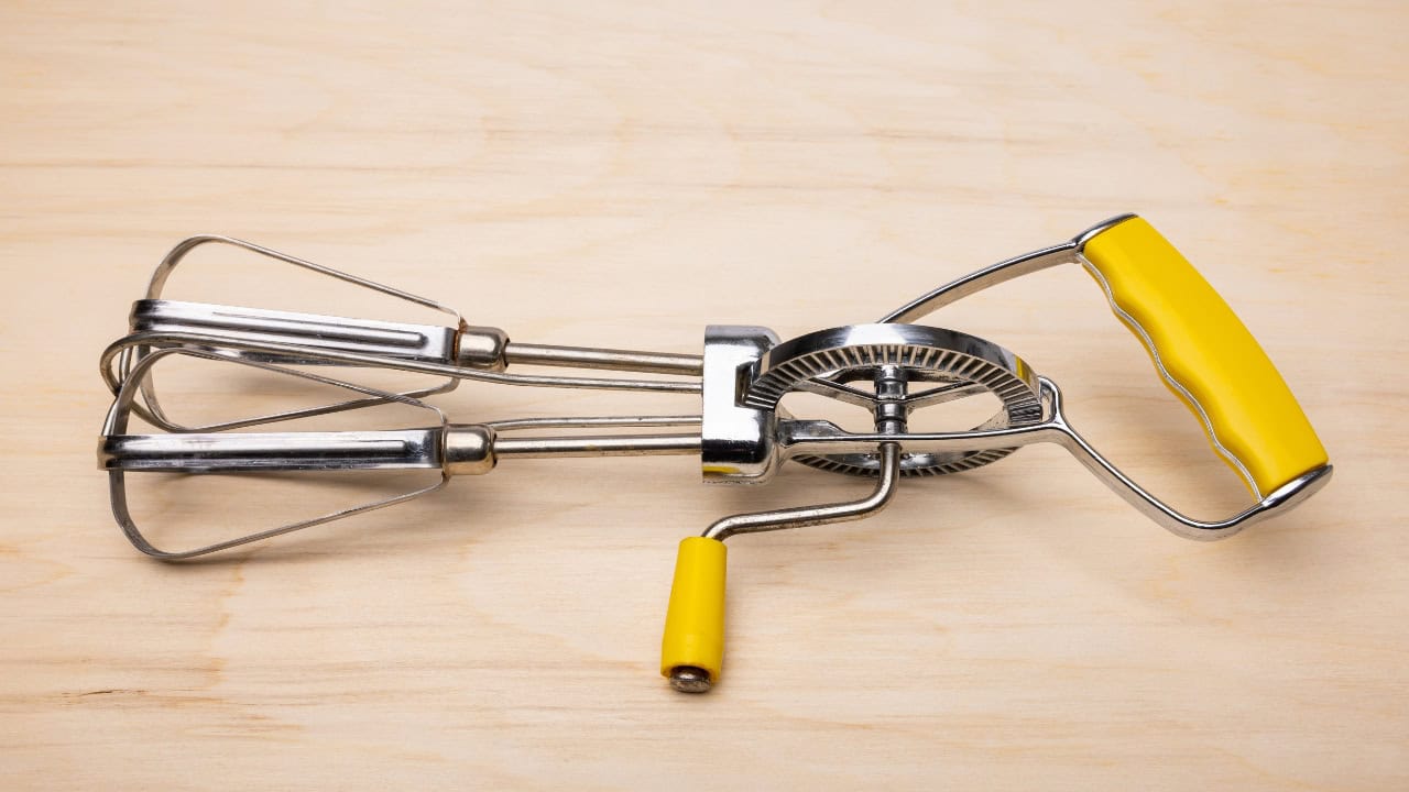 A vintage-style hand-crank egg beater with stainless steel beaters, a yellow handle grip, and a yellow turning knob