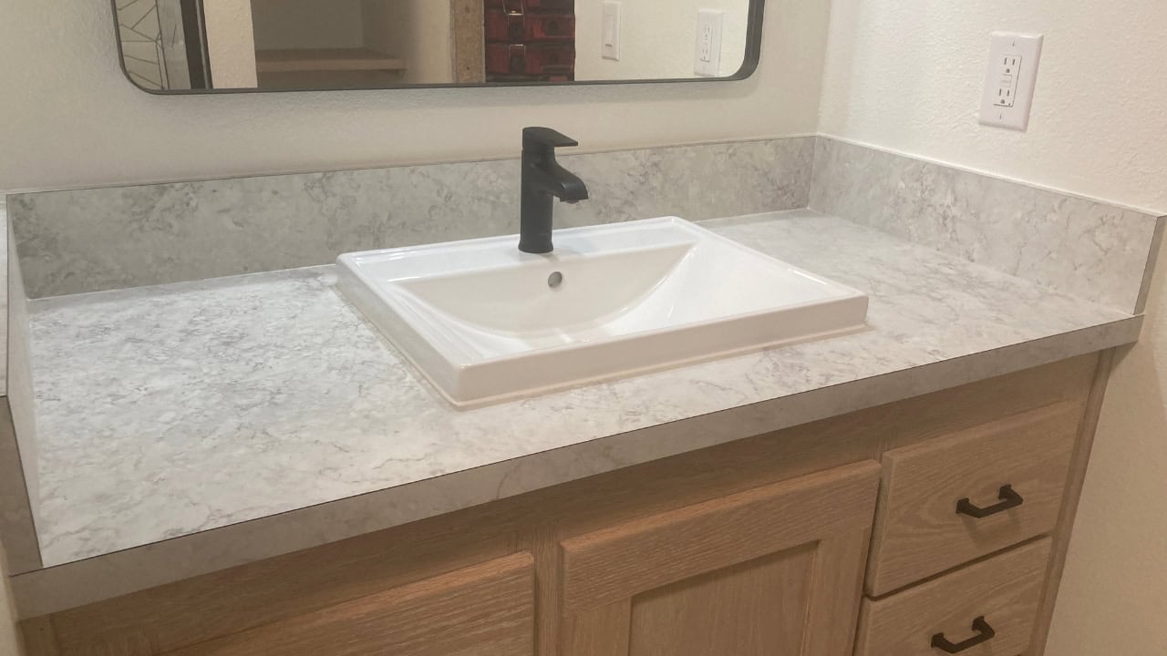 Bathroom vanity with a light gray marble-patterned laminate countertop, rectangular white vessel sink, modern black faucet, wooden cabinet base with drawers, wall-mounted mirror, white backsplash and outlet visible