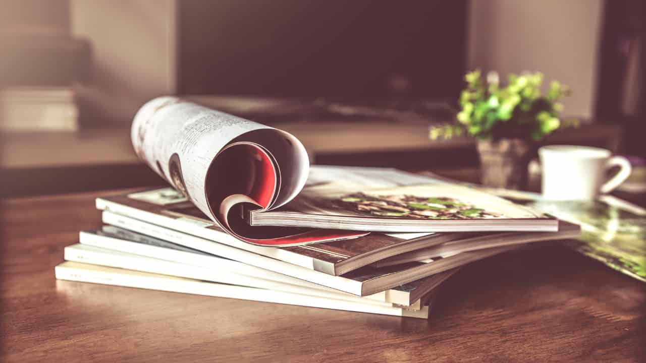 A stack of magazines on a wooden surface, with one magazine partially open, warm lighting, blurred background, cozy and relaxed reading atmosphere