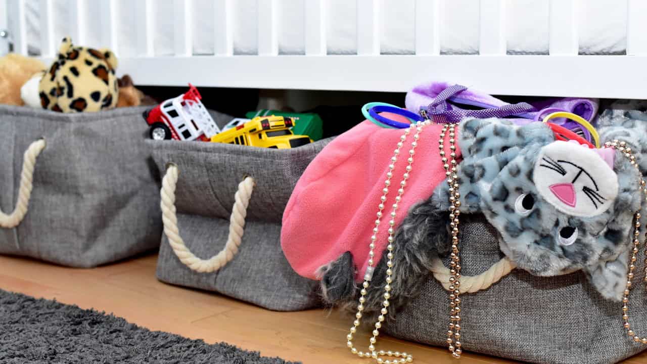 A gray fabric storage basket with rope handles containing various items including a pink hat, pearls, and other accessories on a wooden surface