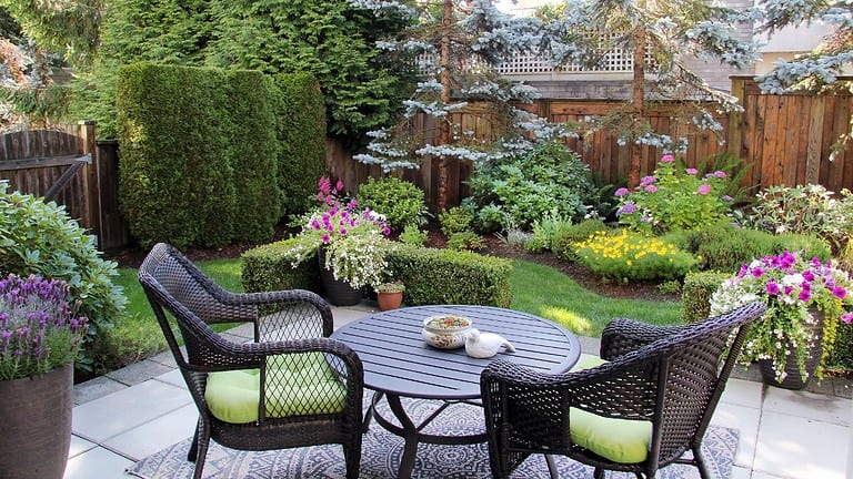 A cozy outdoor seating area with a round table and four wicker chairs, each with green cushions, surrounded by neatly trimmed bushes and vibrant flowers, creating a peaceful garden space, with trees and plants in the background, ideal for relaxation or dining outdoors