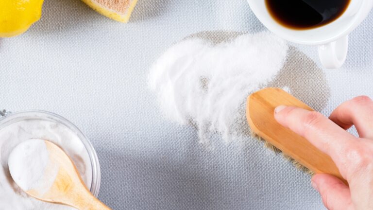 A hand using a brush to scrub baking soda onto a fresh coffee stain on fabric, with a bowl of baking soda, spoon, lemon