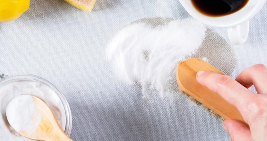 A hand using a brush to scrub baking soda onto a fresh coffee stain on fabric, with a bowl of baking soda, spoon, lemon