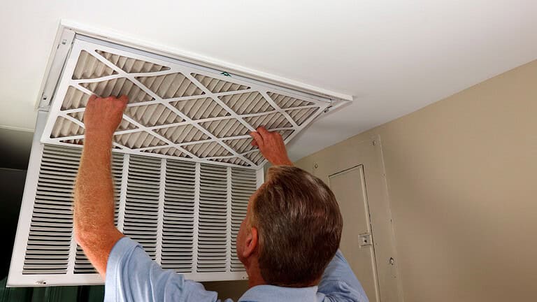 Man in a blue shirt replacing or cleaning an air filter in a ceiling vent, ensuring proper airflow, indoor maintenance