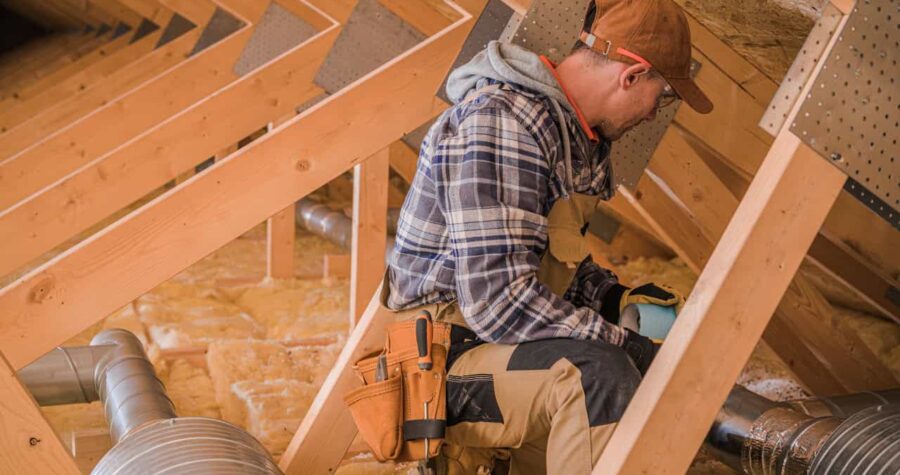 The worker is seated in an attic, installing insulation between wooden beams, conducting attic repairs or improvements, enhance energy efficiency in the home