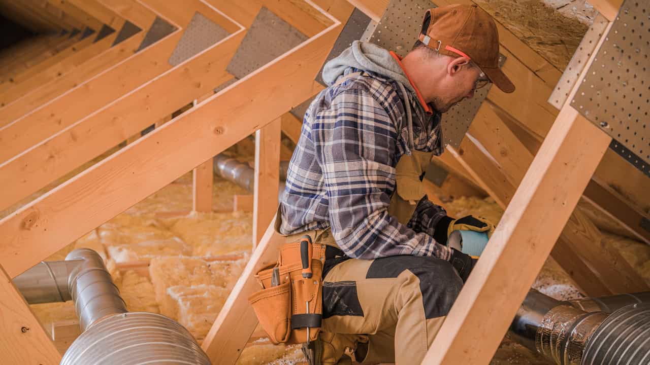 The worker is seated in an attic, installing insulation between wooden beams, conducting attic repairs or improvements, enhance energy efficiency in the home