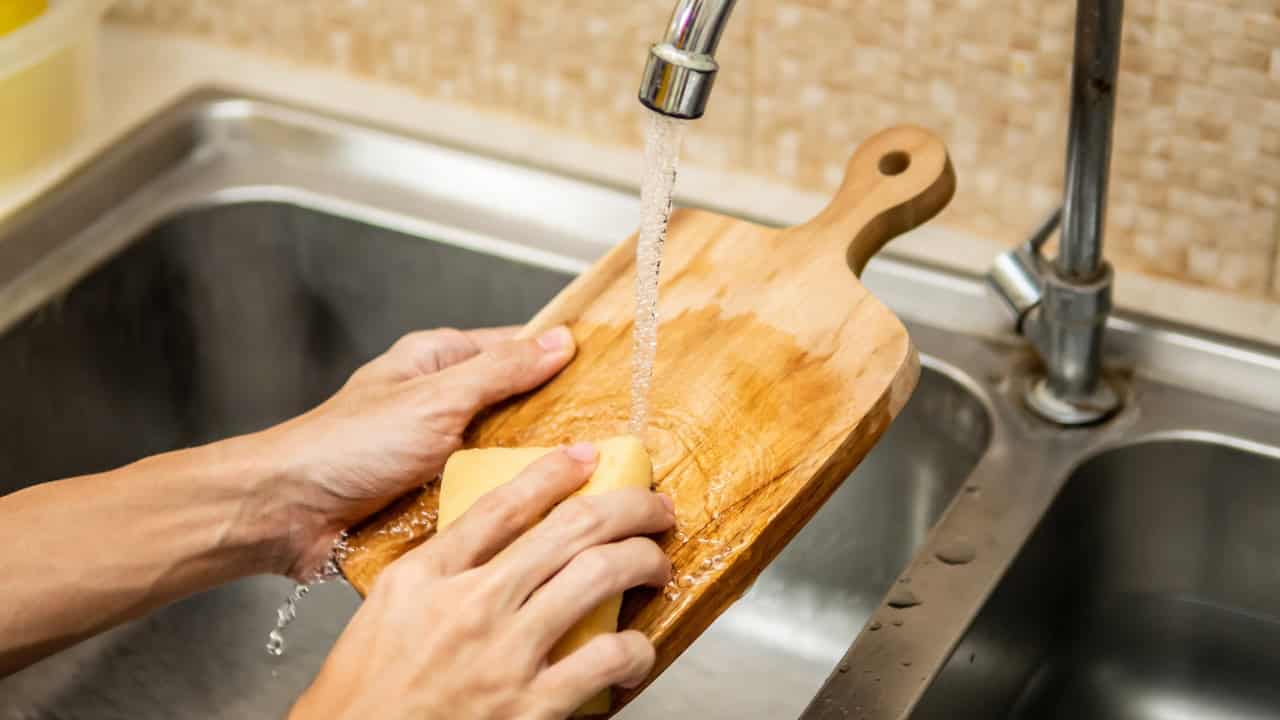 Washing a wooden cutting board, person scrubbing with a yellow sponge, water flowing from faucet, cleaning kitchen utensils, maintaining hygiene