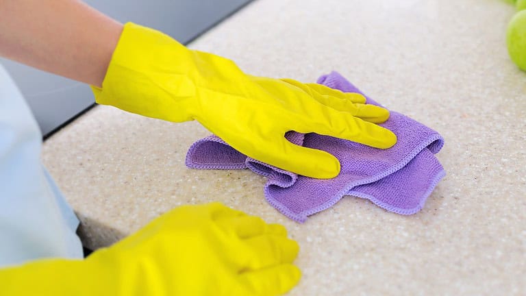 a close-up of two hands wearing yellow rubber gloves cleaning a light-colored countertop with a purple cloth