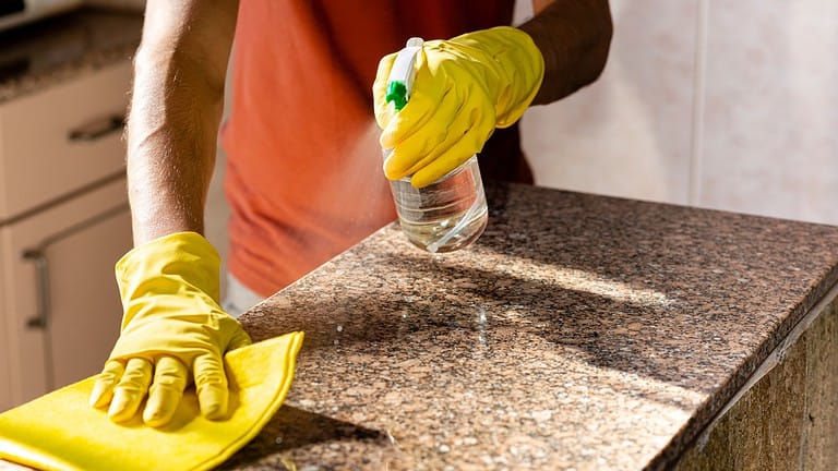 Spraying cleaner on a granite countertop, wearing yellow gloves, cleaning with a cloth, maintaining cleanliness, removing dirt or stains, surface cleaning