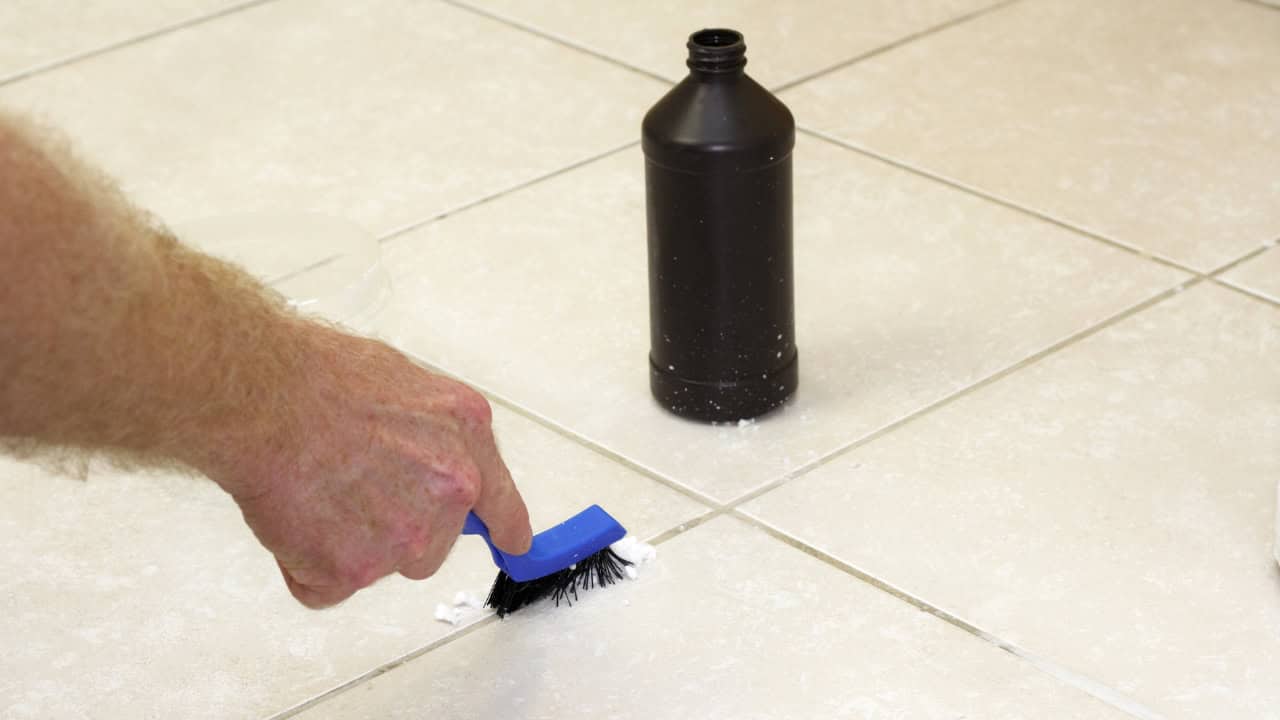 A person scrubbing white grout lines between beige floor tiles with a small blue-handled brush, next to an open black cleaning bottle