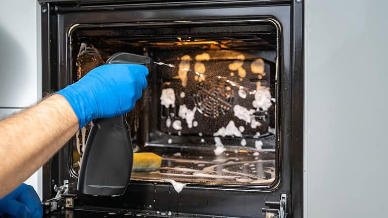 Person wearing blue glove cleaning an oven interior, spray cleaner visible, dirty oven with white cleaning solution splatter, dark oven interior, household cleaning task