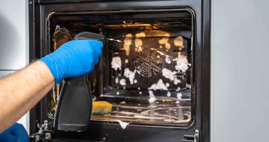 Person wearing blue glove cleaning an oven interior, spray cleaner visible, dirty oven with white cleaning solution splatter, dark oven interior, household cleaning task