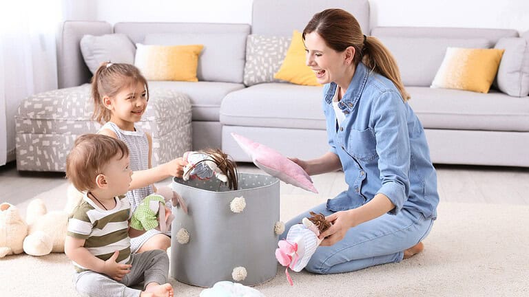 A woman in a blue shirt sits on the floor next to a gray laundry basket, folding or sorting colorful clothes with a couch and pillows visible in the background of a living room