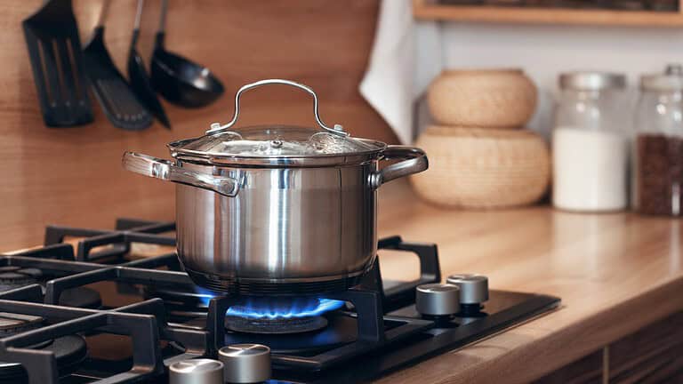 A stainless steel pot with a lid, placed on a gas stove, boiling with steam rising, cooking on a blue flame, in a modern kitchen setting