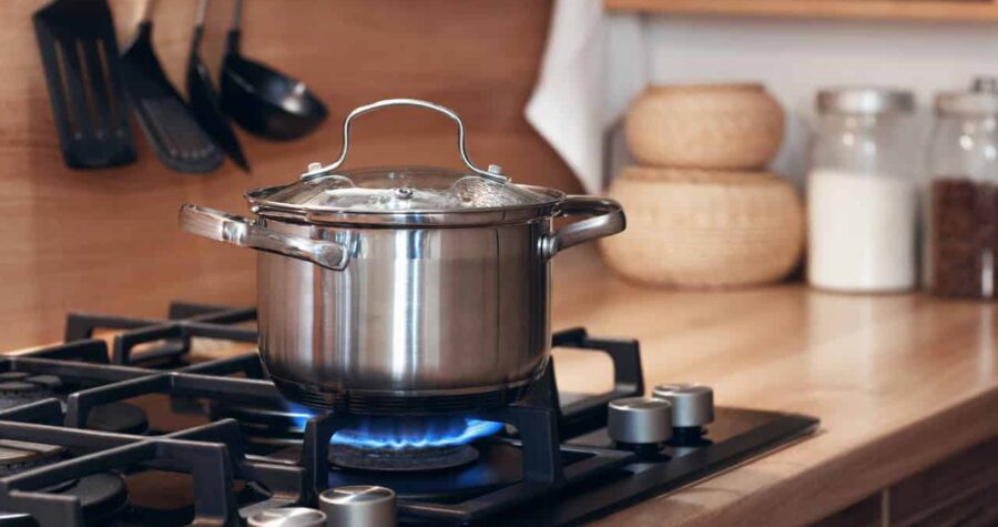 A stainless steel pot with a lid, placed on a gas stove, boiling with steam rising, cooking on a blue flame, in a modern kitchen setting