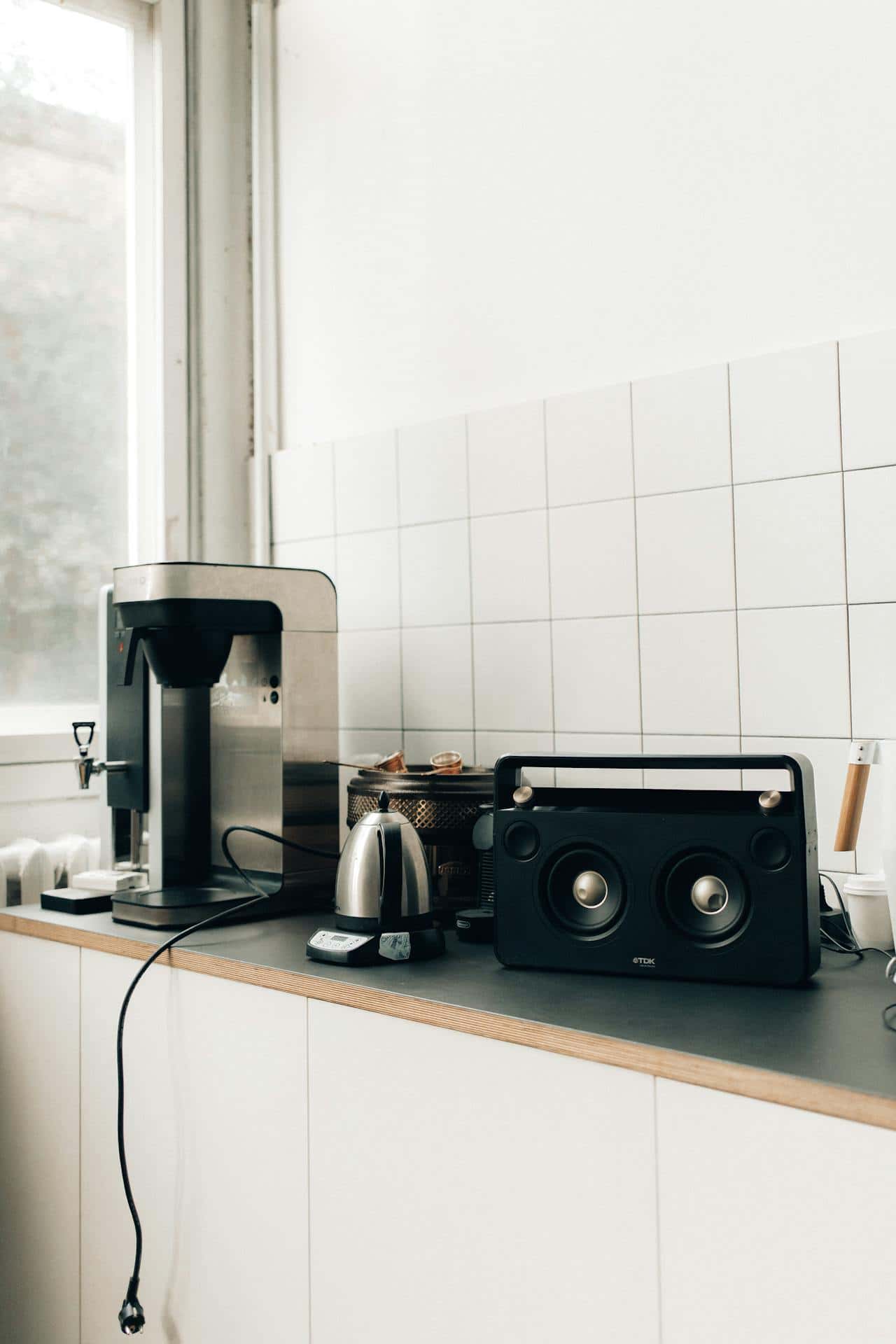 Modern kitchen countertop, stainless steel coffee machine, electric kettle on a digital scale, black portable speaker with gold accents, white tiled wall, window allowing natural light, neatly arranged appliances, minimalistic and clean design.