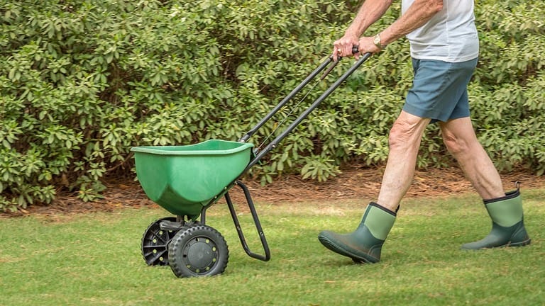 Person pushing a green wheelbarrow, fertilizing lawn, walking on grass, wearing rubber boots, gardening equipment, transporting materials