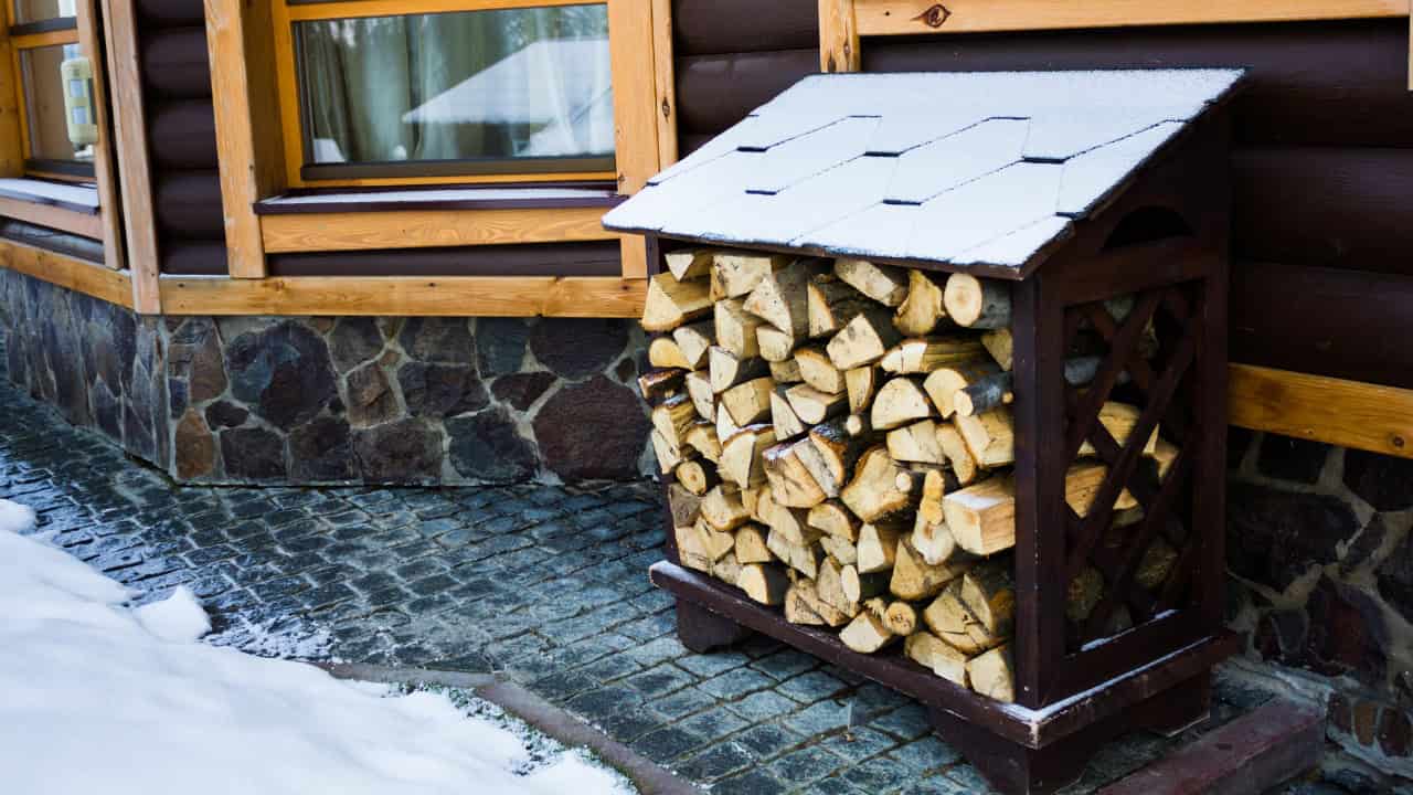 Stacked firewood neatly arranged in a wooden storage rack with a snow-covered slanted roof, placed outside a log cabin with a stone foundation