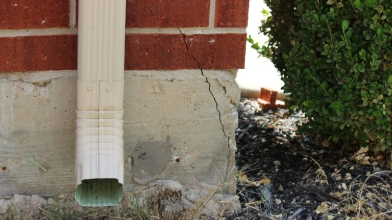 Visible crack in concrete foundation wall near white downspout against red brick house with landscaping