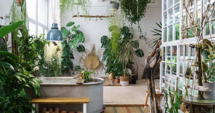 A cozy indoor space with a large metal bathtub, surrounded by various plants in pots, hanging greenery from the ceiling, a rustic wooden shelf, and a natural rug on the floor, creating a peaceful, nature-inspired atmosphere