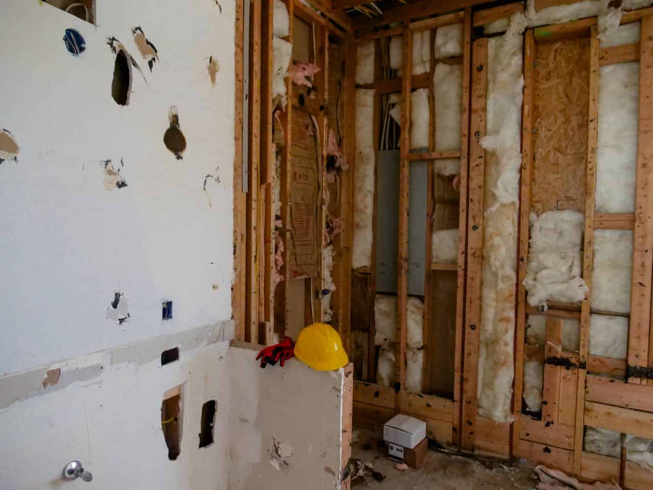 Partially demolished room with exposed wooden framing, insulation, and damaged drywall, with a yellow hard hat and tools on a broken surface