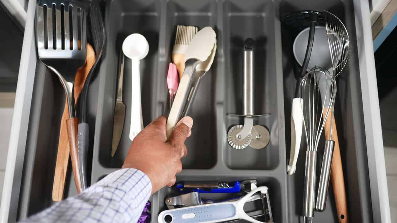 Hand reaching into organized kitchen drawer with compartments containing various utensils, spatulas, whisks, and cooking tools
