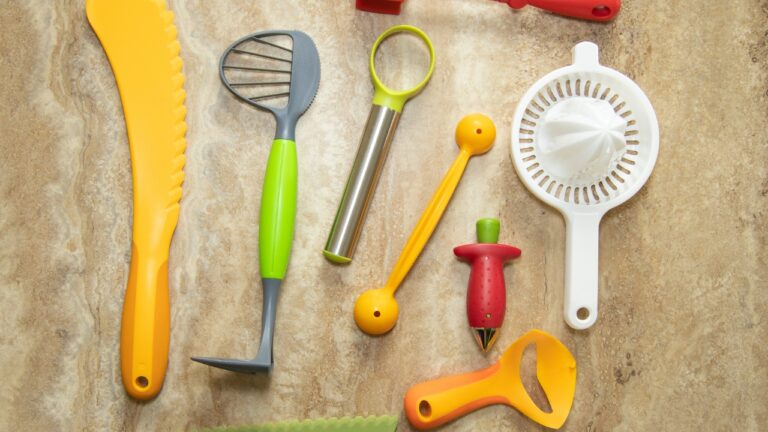 Collection of kitchen utensils on a beige surface, including a lime green whisk, stainless steel tool, orange peeler, red corkscrew, and yellow-handled gadgets. A blue check mark appears in corner