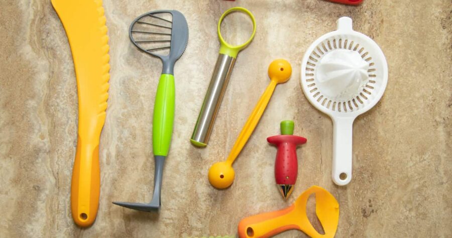 Collection of kitchen utensils on a beige surface, including a lime green whisk, stainless steel tool, orange peeler, red corkscrew, and yellow-handled gadgets. A blue check mark appears in corner