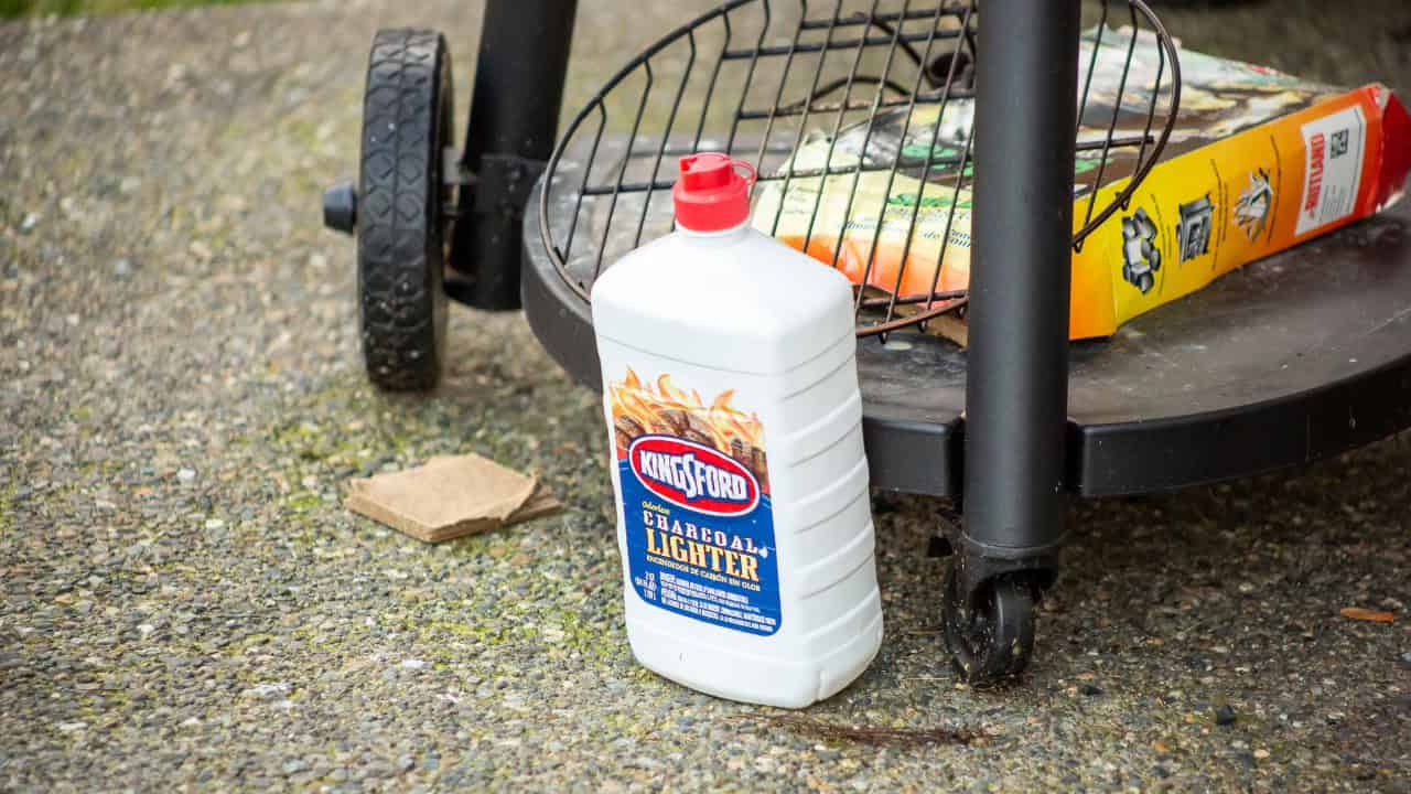 Bottle of charcoal lighter fluid placed on the ground, white plastic container with a red cap, label displaying fire graphics and brand name, positioned next to a black outdoor grill