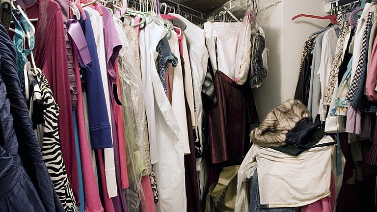 The image shows a closet filled with clothes on hangers, a mix of colorful garments, including dresses and jackets, some folded clothes at the bottom, a cluttered but organized space showcasing a variety of clothing items arranged within a small storage area