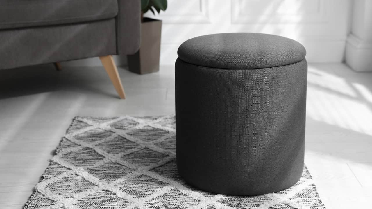 Round dark gray ottoman or pouf on patterned area rug, wooden furniture leg visible in background, minimalist home interior setting