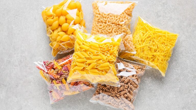 Assorted pasta varieties, packed in clear plastic bags, different shapes and colors, displayed on a light surface, organized for storage