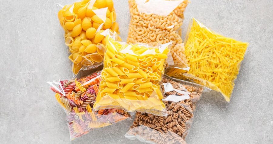 Assorted pasta varieties, packed in clear plastic bags, different shapes and colors, displayed on a light surface, organized for storage