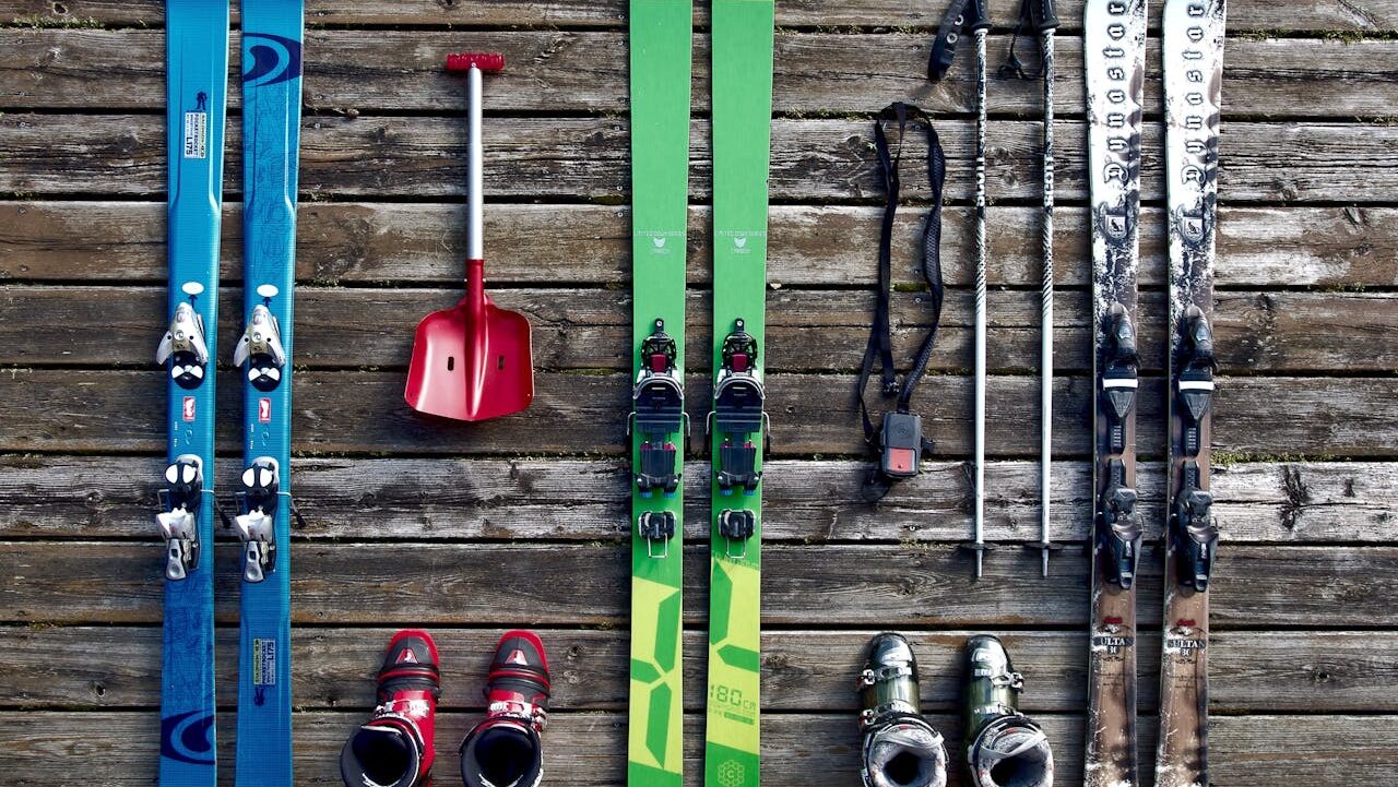 Ski equipment laid out on wooden surface, green and blue skis, ski poles, ski boots, shovel, ski gear preparation, winter sports setup