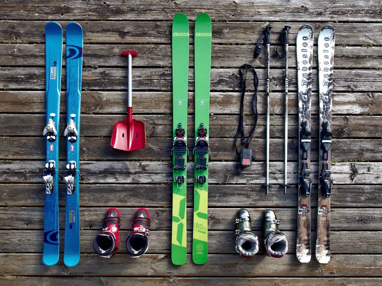 Ski equipment laid out on wooden surface, green and blue skis, ski poles, ski boots, shovel, ski gear preparation, winter sports setup