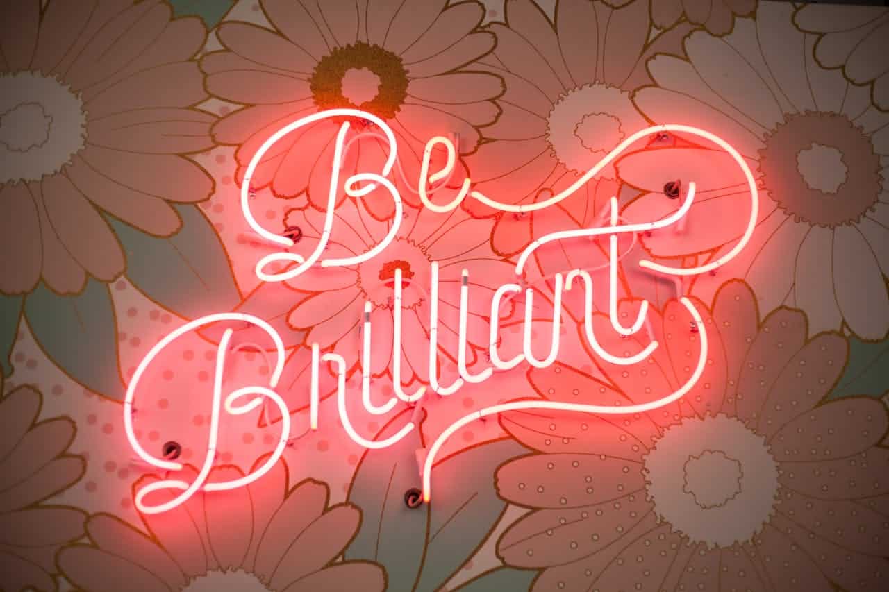 Pink neon sign saying 'Be Brilliant' mounted on a textured wall with floral patterns, soft glow illuminating surrounding area