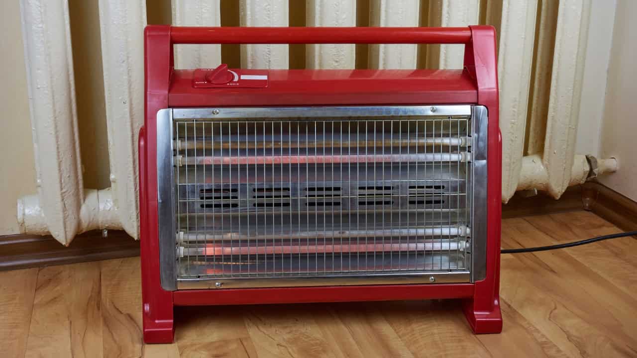 Red portable electric space heater with metal grille, heating elements visible inside, placed on wooden floor, box-shaped design, household heating appliance
