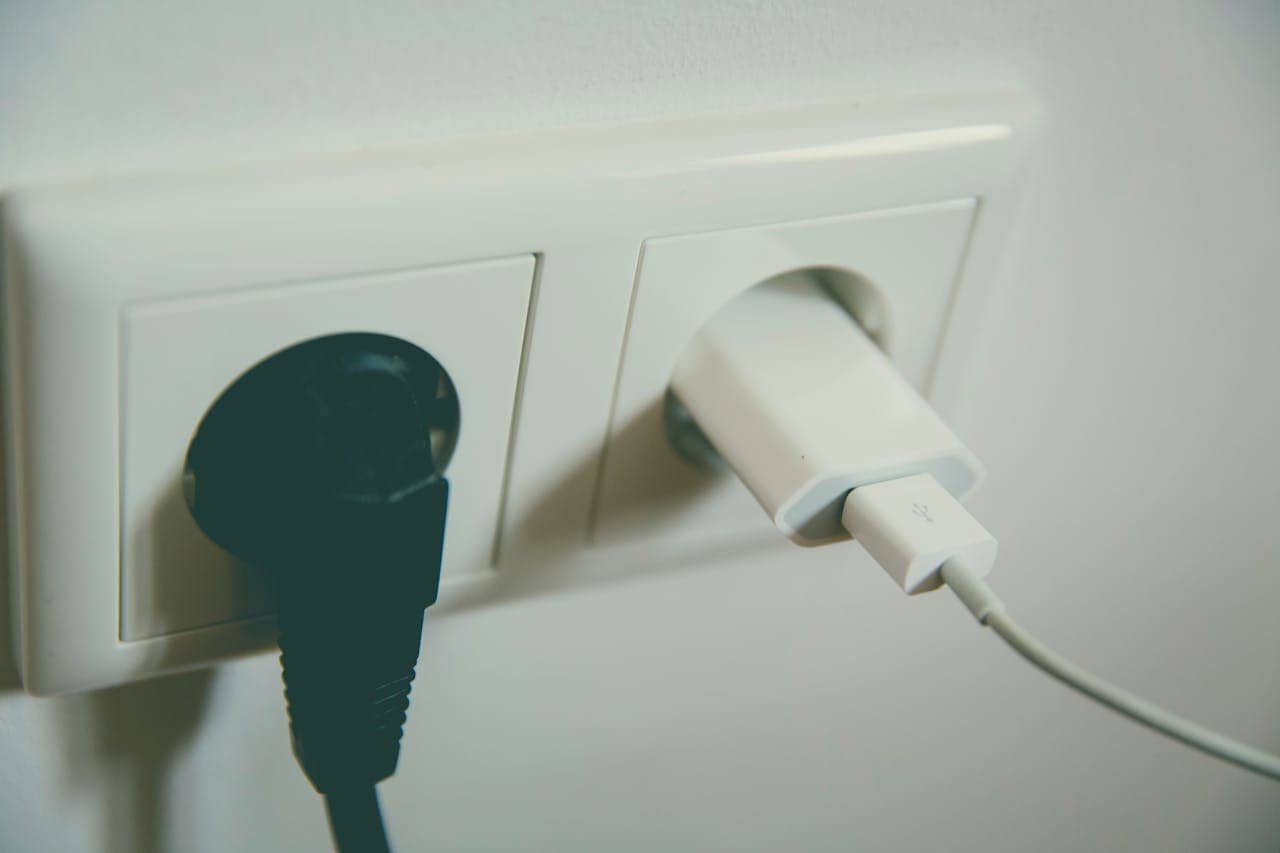 A close-up of a white dual power outlet on a wall, one socket occupied by a black power plug, the other by a white USB charger with a connected cable, minimalistic and modern design, slightly dim lighting.