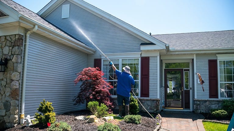 Person in blue shirt washing windows of gray vinyl-sided house, red Japanese maple in landscaped garden bed, sunny day