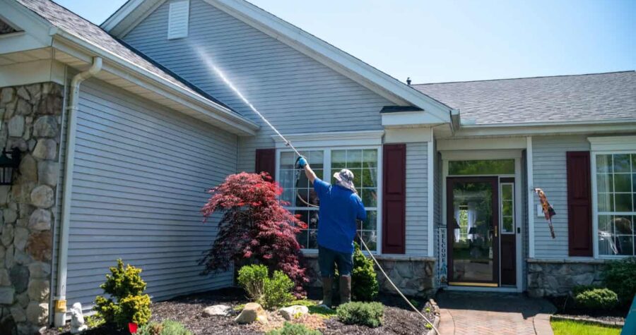 Person in blue shirt washing windows of gray vinyl-sided house, red Japanese maple in landscaped garden bed, sunny day