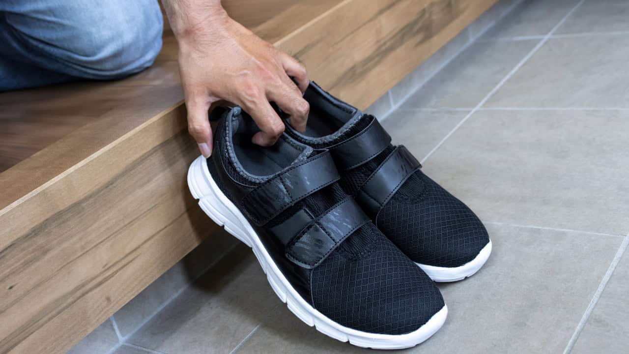 A hand grabbing a pair of black sneakers with Velcro straps, athletic shoes, preparing to wear, shoes placed on a flat surface, comfortable footwear, indoor setting